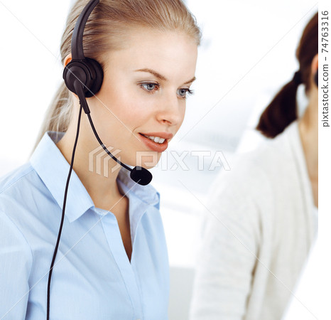 Blond woman call operator is using computer and headset for consulting clients online. Group of diverse people working as customer service occupation. Business concept 74763316
