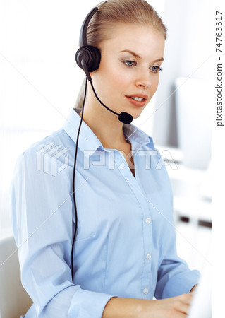 Blond woman call operator is using computer and headset for consulting clients online. Group of diverse people working as customer service occupation. Business concept 74763317