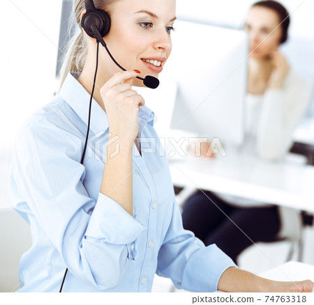 Blond woman call operator is using computer and headset for consulting clients online. Group of diverse people working as customer service occupation. Business concept 74763318