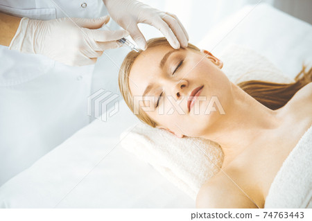 Beautician doctor doing beauty procedure with syringe to blonde female face. Cosmetic medicine, beauty injections concept Beautician doctor doing beauty procedure with syringe to blonde female face. Cosmetic medicine, beauty injections concept 74763443