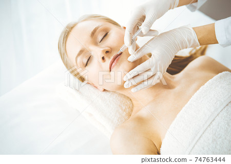 Beautician doctor doing beauty procedure with syringe to blonde female face. Cosmetic medicine, beauty injections concept Beautician doctor doing beauty procedure with syringe to blonde female face. Cosmetic medicine, beauty injections concept 74763444