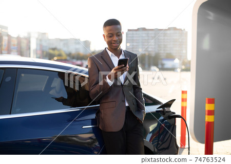 Front view of young happy pleasant African man in treandy formal wear, feeling relaxed, using phone while recharging his modern luxury electric car at the city station Front view of young happy pleasant African man in treandy formal wear, feeling relaxed, using phone while recharging his modern luxury electric car at the city station 74763524