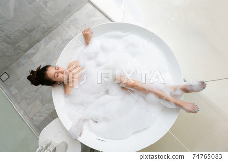 Asian beautiful young woman enjoying pleasant bath with foam, lying with closed eyes. Top view 74765083