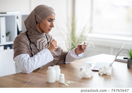 Sick muslim woman doctor looking at thermometer Sick muslim woman doctor looking at thermometer 74765647