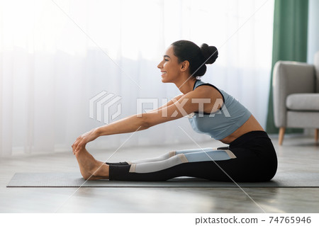 Sporty woman sitting on yoga mat, stretching at home Sporty woman sitting on yoga mat, stretching at home 74765946