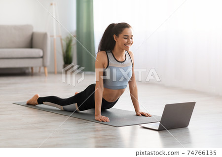 Athletic young woman doing yoga in living room, using laptop Athletic young woman doing yoga in living room, using laptop 74766135