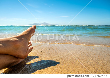 Long slim young woman legs relaxing lying down and sunbathing on sand tropical beach under hot sun in summer. Skincare, sun aging protection and sea travel concept. 74767434