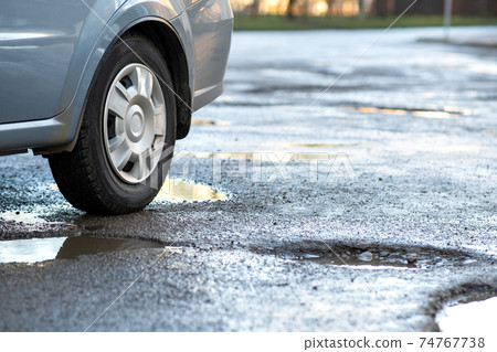 Close up of car wheel on a road in very bad condition with big potholes full of dirty rain water pools. Close up of car wheel on a road in very bad condition with big potholes full of dirty rain water pools. 74767738