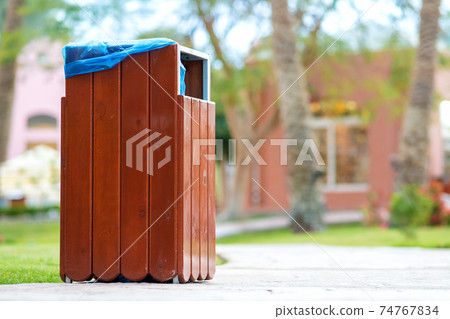 Yellow wooden trash can outdoors on the side of sidewalk in park. Garbage container outside. 74767834