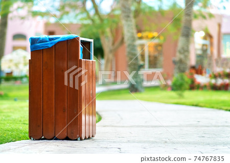 Yellow wooden trash can outdoors on the side of sidewalk in park. Garbage container outside. 74767835