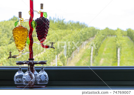 glass carafe with white and red wine in the shape of a grape and vineyard background glass carafe with white and red wine in the shape of a grape and vineyard background 74769007