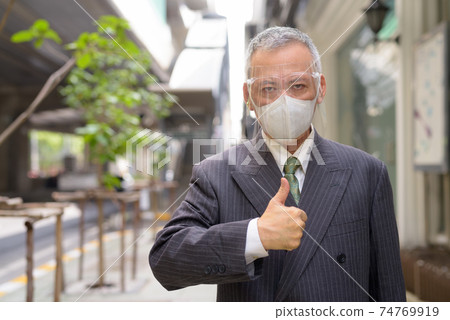 Mature Japanese businessman with mask and face shield giving thumbs up in the city Mature Japanese businessman with mask and face shield giving thumbs up in the city 74769919