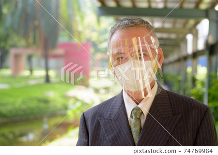Mature Japanese businessman wearing mask and face shield at the park Mature Japanese businessman wearing mask and face shield at the park 74769984