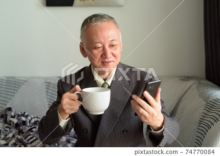 Happy mature Japanese businessman using phone while drinking coffee at home Happy mature Japanese businessman using phone while drinking coffee at home 74770084