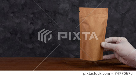 Pouch bag mockup banner. Hand in protective gloves holds brown kraft paper pack with coffee beans wooden background. 74770673