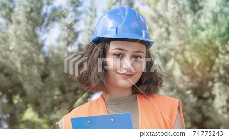 Portrait of a cheerful young woman engineer or worker in a uniform and protective blue hardhat, smiling and looking at camera at construction site Portrait of a cheerful young woman engineer or worker in a uniform and protective blue hardhat, smiling and looking at camera at construction site 74775243