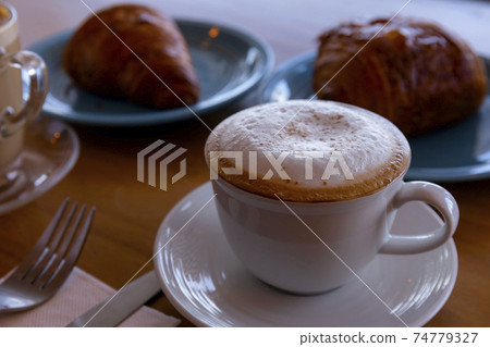Close up hot cappuccino, latte, caramel macchiato on table with blur coffee shop background. Hot cappuccino coffee on wood table with blur croissant background. breakfast at coffee shop. softfocus Close up hot cappuccino, latte, caramel macchiato on table with blur coffee shop background. Hot cappuccino coffee on wood table with blur croissant background. breakfast at coffee shop. softfocus 74779327