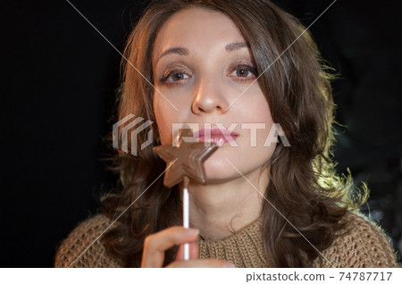 Female portrait of a girl holding chocolate star on stick and looking at the camera on black background in studio. Diet and food concept Female portrait of a girl holding chocolate star on stick and looking at the camera on black background in studio. Diet and food concept 74787717