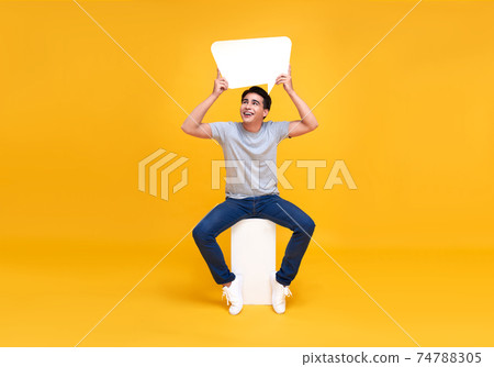 Asian man sitting and holding blank speech bubbles. looking up with smile on yellow background. 74788305