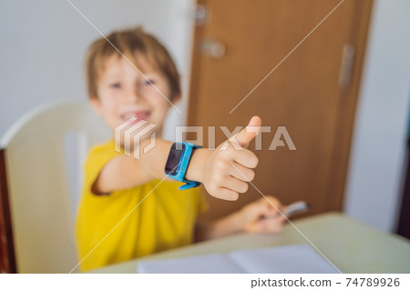 Little boy sitting at the table and looking smart watch. Smart watch for baby safety. The child makes school lessons, listening to music, calling friends Little boy sitting at the table and looking smart watch. Smart watch for baby safety. The child makes school lessons, listening to music, calling friends 74789926