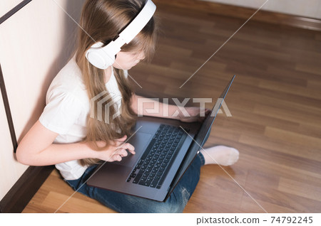 A little girl in white headphones with a laptop on her lap sits on the floor. look with a serious face in your laptop A little girl in white headphones with a laptop on her lap sits on the floor. look with a serious face in your laptop 74792245