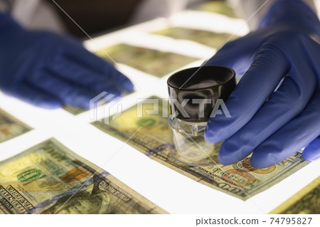 Man in gloves checking counterfeit money with magnifying glass closeup 74795827