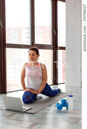 Home online workout. Middle aged woman stretching legs at home in front of laptop monitor. Vertical photo. Home online workout. Middle aged woman stretching legs at home in front of laptop monitor. Vertical photo. 74796027