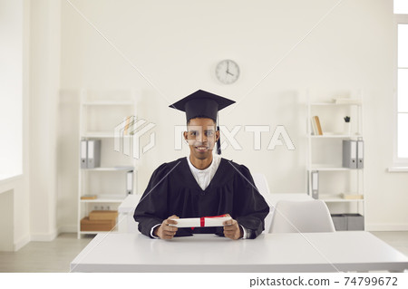 Happy black boy student university graduate in mantle and bonet sitting at desk with diploma Happy black boy student university graduate in mantle and bonet sitting at desk with diploma 74799672