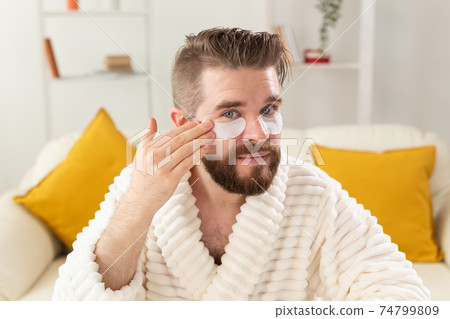 Bearded man sitting with eye patches on his face. Wrinkles and face home care for men. Bearded man sitting with eye patches on his face. Wrinkles and face home care for men. 74799809