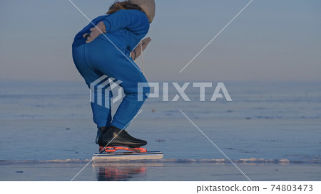 The girl train on ice speed skating. The child skates in the winter in blue sportswear suit, sport glasses. Children speed skating sport. The girl train on ice speed skating. The child skates in the winter in blue sportswear suit, sport glasses. Children speed skating sport. 74803473