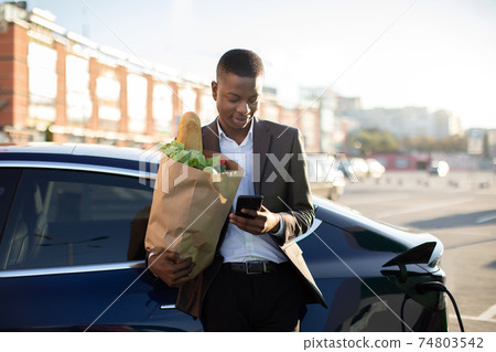 Urban, electric vehicle, eco concept. Young black skinned businessman in stylish suit, browsing on cellphone while waiting for his electric car charging, holding big bag with fresh food after shopping 74803542