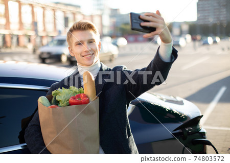 Portrait of young handsome european man, making selfie photo on his smartphone while leaning on his electric car, charging the battery at city power station after buying frsh products in trade mall Portrait of young handsome european man, making selfie photo on his smartphone while leaning on his electric car, charging the battery at city power station after buying frsh products in trade mall 74803572