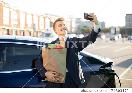 Cheerful laughing young guy standing with shopping bag with vegetables near new modern charging electric car with smartphone in hands and having video call Cheerful laughing young guy standing with shopping bag with vegetables near new modern charging electric car with smartphone in hands and having video call 74803573