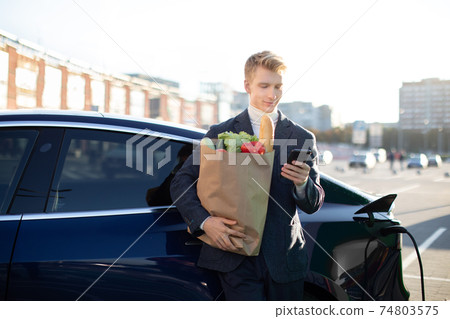 Handsome young smiling Caucasian man, holding shopping bag with vegetables, messaging on his mobile phone or using app for paying, while charging electric car outdoors at city refueling station Handsome young smiling Caucasian man, holding shopping bag with vegetables, messaging on his mobile phone or using app for paying, while charging electric car outdoors at city refueling station 74803575