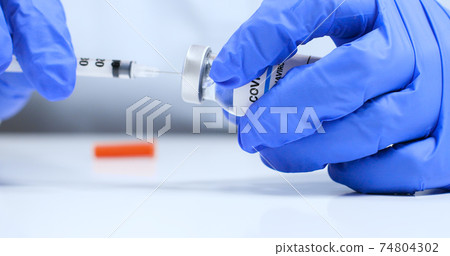 Female doctor hands in blue medical gloves with transparent liquid coronavirus vaccine bottle and syringe for injection in the laboratory. Research and development of new cure for diseases 74804302