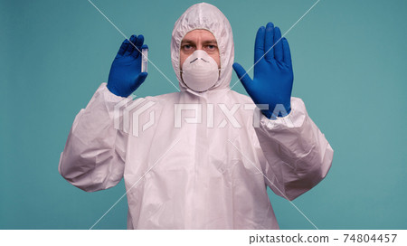 A male doctor in protective overalls and a mask holds an ampoule with a vaccine in his hands. A male doctor in protective overalls and a mask holds an ampoule with a vaccine in his hands. 74804457