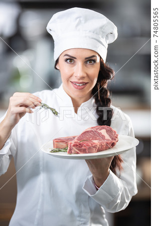 Beautiful female chef in uniform and hat holds a plate with raw steaks and fresh herbs in the kitchen Beautiful female chef in uniform and hat holds a plate with raw steaks and fresh herbs in the kitchen 74805565