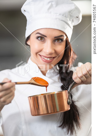 Professional female chef holds a wooden spoon with orange sauce and a pan wearing hat and chef's uniform Professional female chef holds a wooden spoon with orange sauce and a pan wearing hat and chef's uniform 74805567
