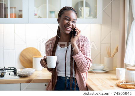 Cheerful black woman talking on mobile phone and drinking coffee in kitchen 74805945