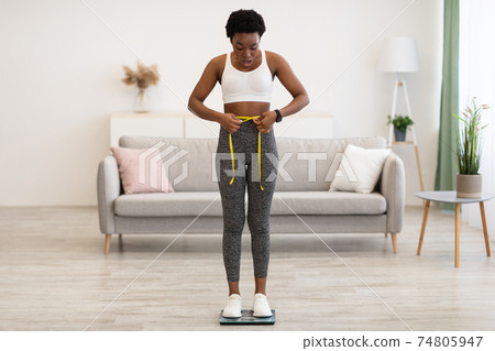 Shocked Black Woman Weighing Herself And Measuring Waist At Home Shocked Black Woman Weighing Herself And Measuring Waist At Home 74805947