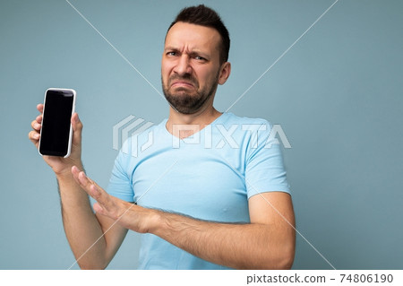 Dissatisfied handsome young unshaven brunet man wearing everyday blue t-shirt isolated over blue 74806190