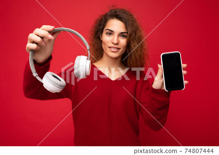 Charming funny young brunette curly woman wearing dark red sweater isolated on red backdrop holding 74807444