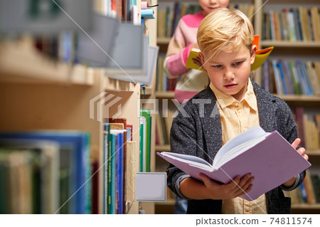 boy stands reading a book against multi colored bookshelf in library 74811574