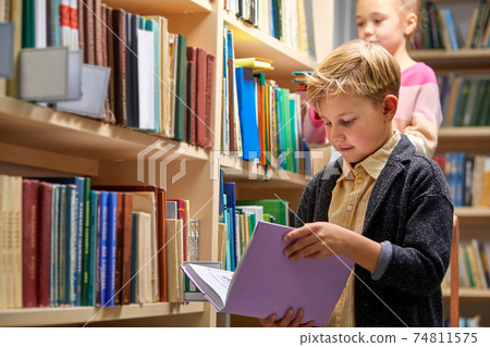 boy stands reading a book against multi colored bookshelf in library 74811575