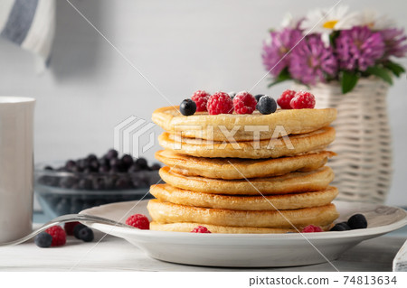 Stack of pancakes with fresh berries on a white plate and a vase of wildflowers on the table Stack of pancakes with fresh berries on a white plate and a vase of wildflowers on the table 74813634