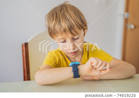 Little boy sitting at the table and looking smart watch. Smart watch for baby safety. The child makes school lessons, listening to music, calling friends Little boy sitting at the table and looking smart watch. Smart watch for baby safety. The child makes school lessons, listening to music, calling friends 74819228