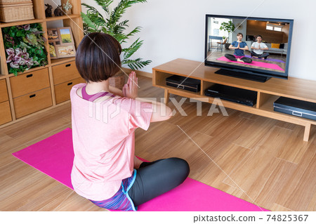 在線瑜伽一個年輕女子在客廳裡看電視屏幕時做瑜伽合掌姿勢 在線瑜伽一個年輕女子在客廳裡看電視屏幕時做瑜伽合掌姿勢 74825367
