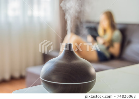 Aromatherapy Concept. Wooden Electric Ultrasonic Essential Oil Aroma Diffuser and Humidifier. Ultrasonic aroma diffuser for home. Woman resting at home Aromatherapy Concept. Wooden Electric Ultrasonic Essential Oil Aroma Diffuser and Humidifier. Ultrasonic aroma diffuser for home. Woman resting at home 74826068