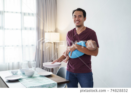 father doing chores at home while carrying his baby 74828832