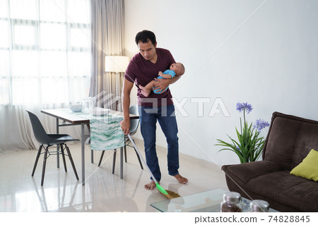 father cleaning the floor using broom while carrying his infant baby 74828845